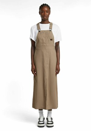 Braunes Baumwolle-Overallkleid mit verstellbaren Trägern, Fronttasche und seitlichem Schlitz. Getragen über einem weißen T-Shirt und kombiniert mit schwarzen Sneakers und Socken.
