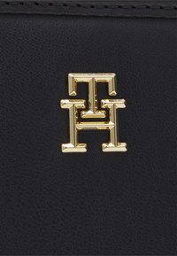 Materiale in pelle nera con un logo metallico dorato che presenta un design geometrico costituito da lettere intrecciate, aggiungendo texture e dettaglio.