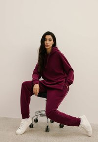 Burgundowa bluza z kapturem i dresy wykonane z miękkiego materiału, o luźnym kroju, z przednią kieszenią, połączone z białymi trampkami na stołku.