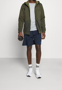 Sweat à capuche vert olive avec une fermeture éclair à l'avant, t-shirt gris, short marine, baskets blanches et bouteille d'eau noire. Souligné par des chaussettes rayées.