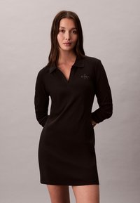 Femme aux longs cheveux bruns portant une robe noire à manches longues avec col, arborant le logo Calvin Klein sur la poitrine, debout devant un fond neutre.