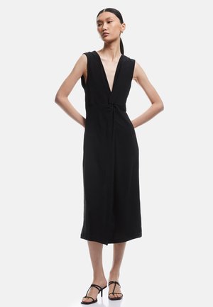 Vrouw staat met de handen in de heupen, draagt een mouwloze zwarte midi-jurk met diepe V-hals en zwarte bandjes met hoge hakken.