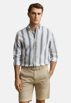 Jonge man die een wit overhemd draagt met opgerolde mouwen en blauwe verticale strepen, en een beige riem met bijpassende korte broek, handen ontspannen in de zakken.