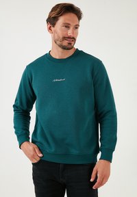 Zöldeskék crewneck pulóver, texturált anyagból, fehér logó hímzéssel a mellkasán, bordás mandzsettákkal és aljjal.