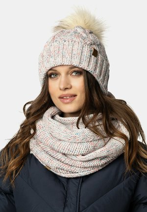 Strik beanie med et lyst pink og hvidt mønster, der har en fluffy pom-pom. Matchende tekstureret tørklæde draperet om halsen.