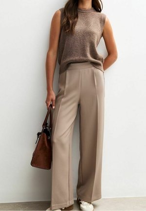 Vrouw draagt een mouwloos bruin gebreid topje, beige broek met wijde pijpen, witte sneakers, houdt een bruine handtas vast, staat tegen een witte muur.