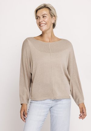 Smiling woman with short blonde hair wears beige long-sleeve sweater and light blue jeans, standing with arms relaxed on a neutral background.