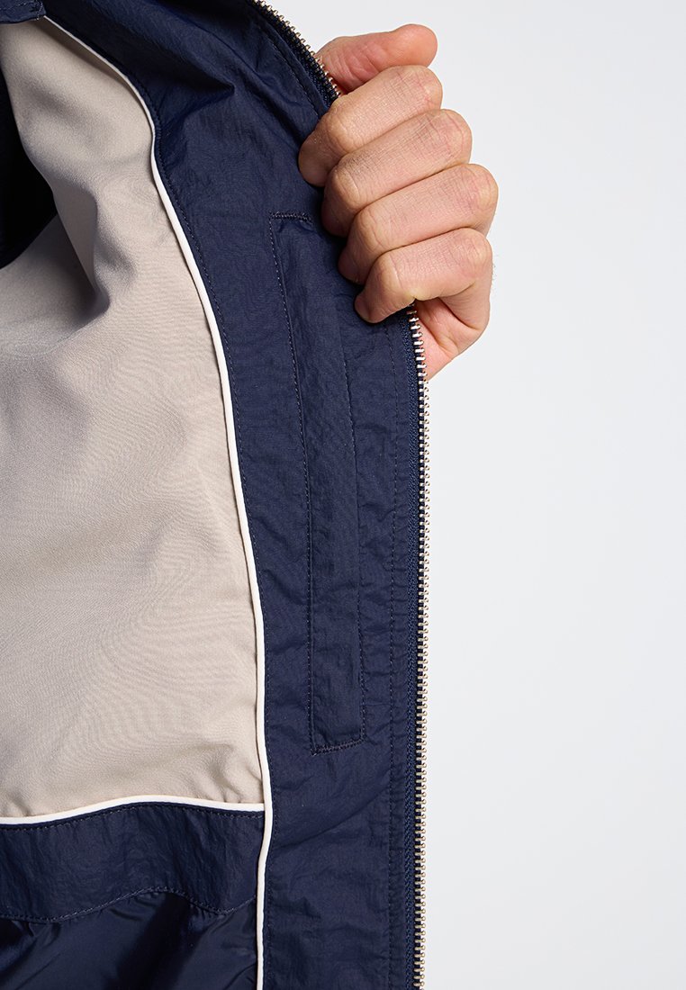 Marineblaue Jacke mit glattem beigem Innenfutter, ausgestattet mit einem Reißverschluss und einer seitlichen Tasche, die in das Futter eingenäht ist.