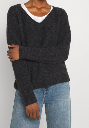 Personne portant un pull gris foncé en matière pelucheuse à col en V par-dessus un haut blanc, assorti à un jean en denim bleu clair, bras croisés sur la poitrine.