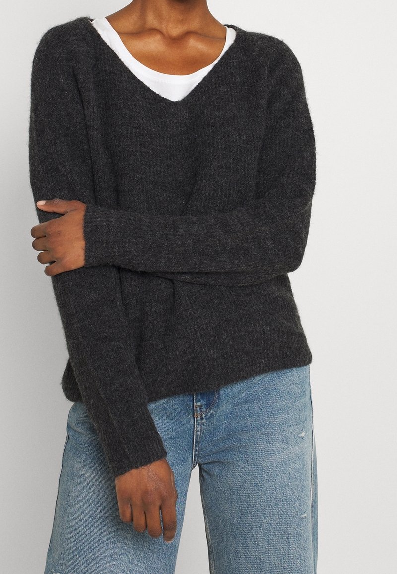 Personne portant un pull gris foncé en matière pelucheuse à col en V par-dessus un haut blanc, assorti à un jean en denim bleu clair, bras croisés sur la poitrine.