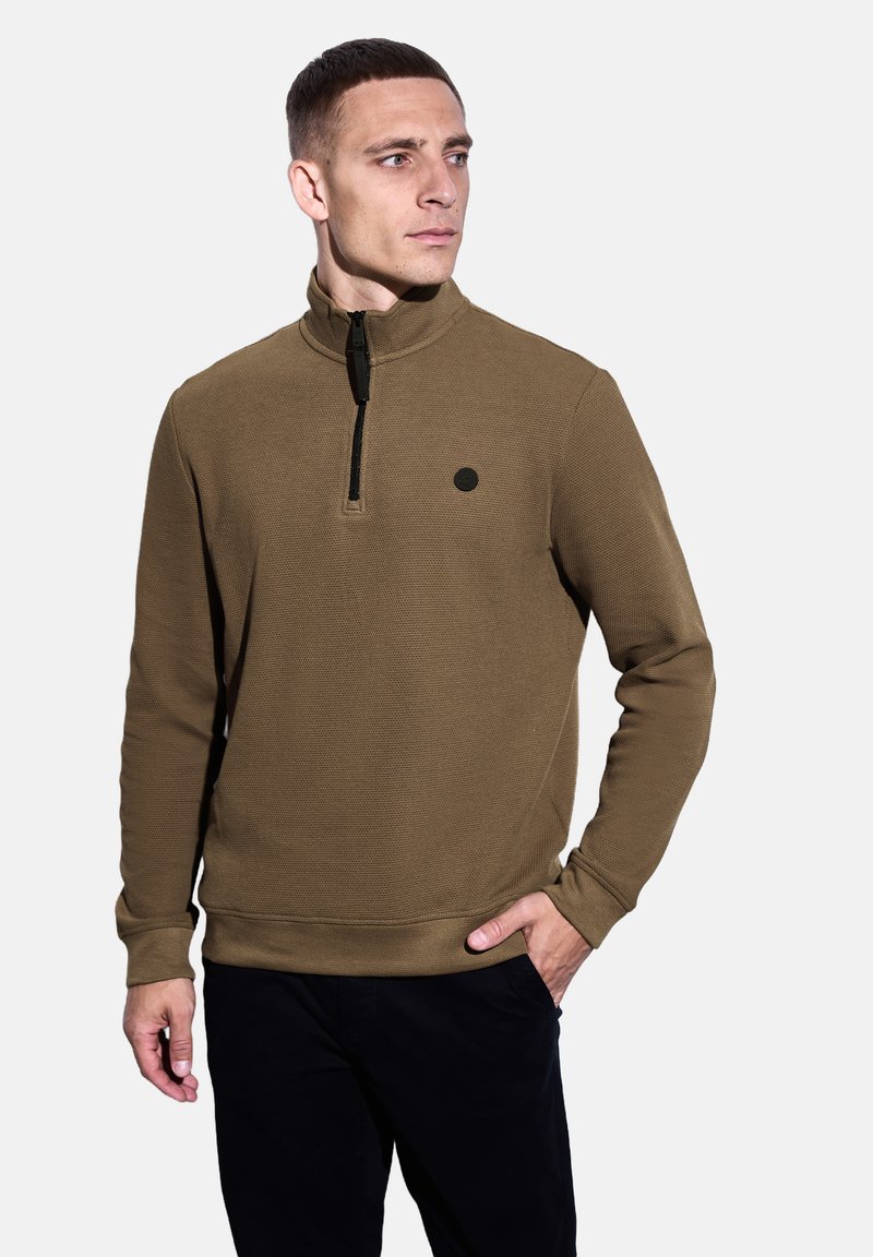 Muž v hnědém dlouhém svršku s krátkým zipem a malým černým logem na hrudi, s černými kalhotami, dívající se doprava.