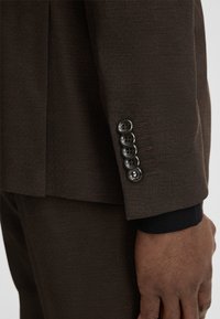 Brauner Anzugsakkoärmel mit strukturiertem Stoff, fünf dunklen Knöpfen und einem gerippten schwarzen Bündchen mit feiner Naht.