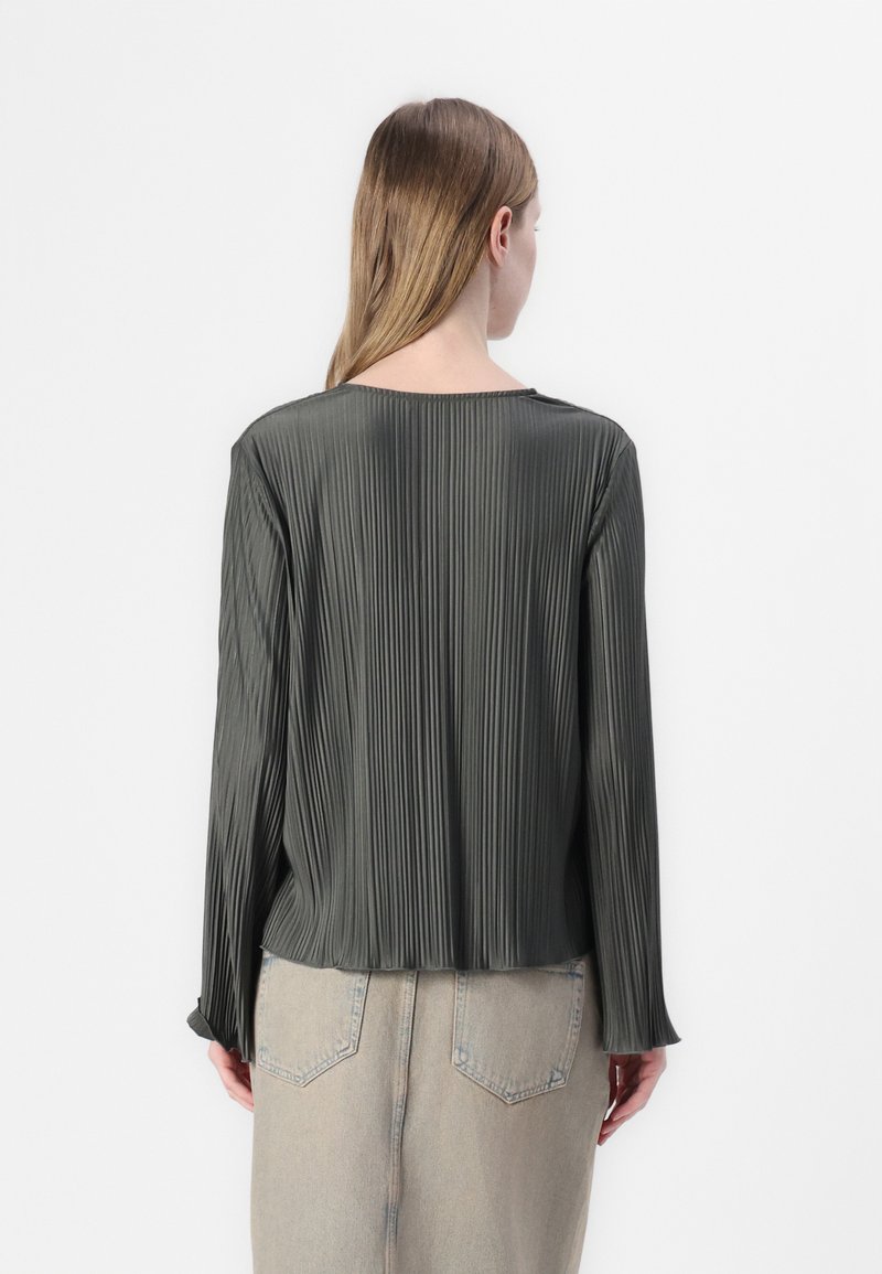 Faltige, dunkelgrüne Bluse mit langen Ärmeln, glatter Textur, rundem Ausschnitt und lockerer Passform. Trägt eine hellfarbige Jeansrock.