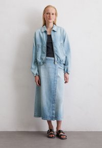Eine junge Frau steht gegen eine weiße Wand und trägt eine hellblaue Jeansjacke und einen passenden Rock, kombiniert mit schwarzen Sandalen.