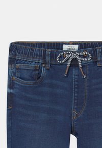 Tmavě modré džíny z denimu s elastickým pasem, stahovací šňůrkou a detaily předních kapes. Šité akcenty v kontrastní barvě.