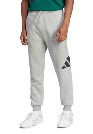Persona con pantalones de chándal Adidas gris claro con cintura ajustable mediante cordón y zapatillas blancas, sobre un fondo blanco.