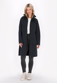 Cappotto di pile blu navy con colletto alto, zip frontale e due tasche laterali; abbinato a pantaloni grigi e sneakers beige. Tessuto strutturato.