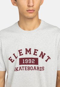 Šedé bavlněné tričko s kulatým výstřihem. Obsahuje vínový text: "ELEMENT 1992 SKATEBOARDS" v tučném písmu. Krátké rukávy.