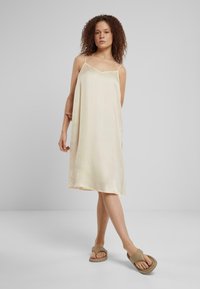 Satin klänning i blek gul med tunna axelremmar, A-linjeformad och sidofickor. Buren med beige sandaler mot en neutral bakgrund.