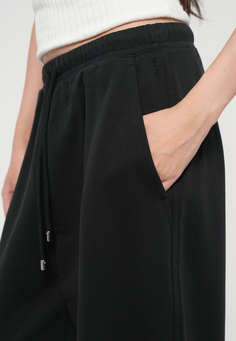 Pantalons de jogging noirs en tissu doux, avec une taille élastique, des cordons de serrage réglables et des poches latérales. Texture lisse et coupe décontractée.