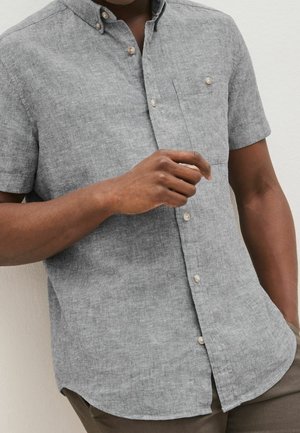 Homme portant une chemise grise à manches courtes avec une poche poitrine et des boutons beiges, debout devant un fond uni.