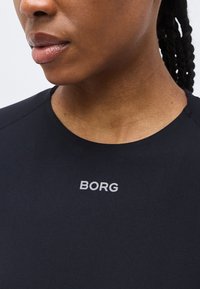 Κοντινή λήψη ενός ατόμου που φοράει μαύρο πουκάμισο με τη λέξη "BORG" τυπωμένη σε γκρι στο στήθος, εμφανίζοντας τα χείλη, τη γραμμή του σαγονιού και πλεξούδες στα μαλλιά.