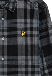 Camisa de franela a cuadros con rayas negras, grises y azules. Destacado logo de águila amarilla en el lado izquierdo del pecho, diseño de botones.