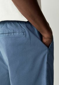 Pantalones cortos de cintura elástica azul con una textura de algodón suave, que cuentan con un bolsillo trasero y un corte relajado. Bolsillo lateral visible en el lado izquierdo.