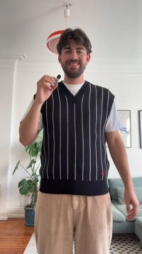 Μαύρο και άσπρο ριγέ γιλέκο με σχέδιο V-neck, κατασκευασμένο από πλεκτό ύφασμα. Φορεμένο πάνω από ένα λευκό πουκάμισο, με μπεζ καμπαρντίνες παντελόνες.