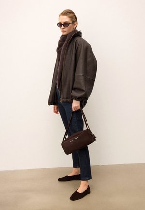 Veste en cuir marron de type bombardier avec une fermeture éclair, associée à un jean foncé et des ballerines en suède marron. Le modèle tient un sac à main zippé marron.