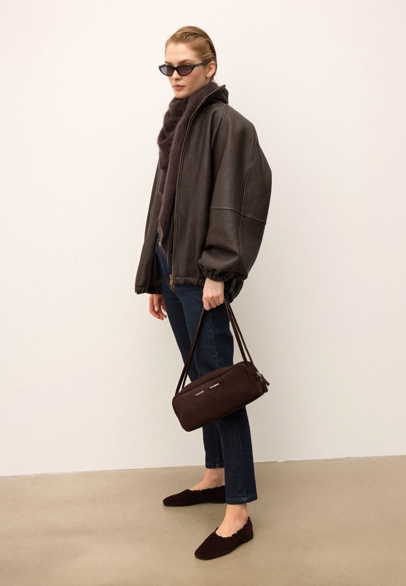 Veste en cuir marron de type bombardier avec une fermeture éclair, associée à un jean foncé et des ballerines en suède marron. Le modèle tient un sac à main zippé marron.