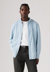 Cămașă din denim de colorare albastru deschis, cu nasturi, cu o croială lejeră, purtată peste un tricou alb. Pantaloni verzi închiși cu o curea neagră, design minimal și fără modele.