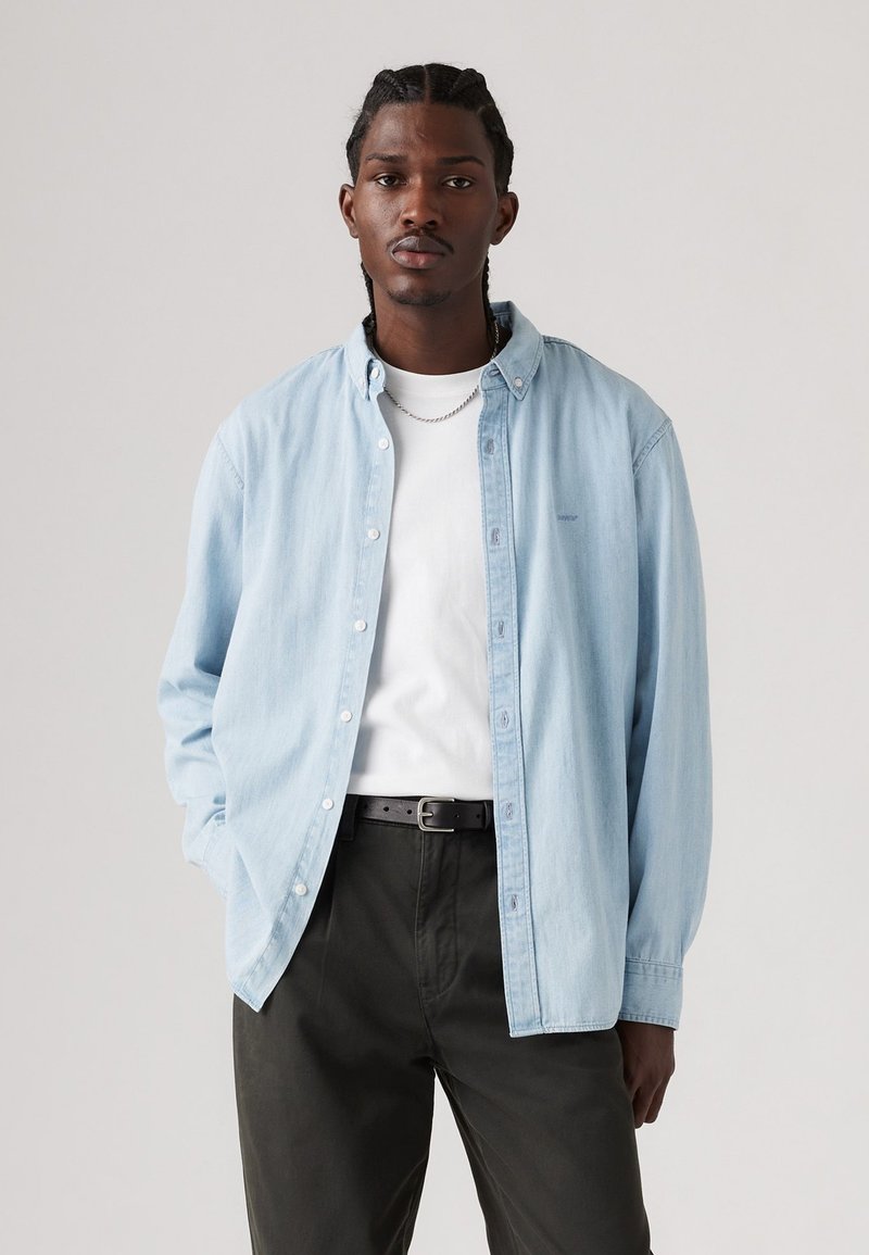 Chemise en denim bleu clair à boutons, coupe décontractée, superposée sur un t-shirt blanc. Pantalons vert foncé avec une ceinture noire, design minimaliste et sans motifs.