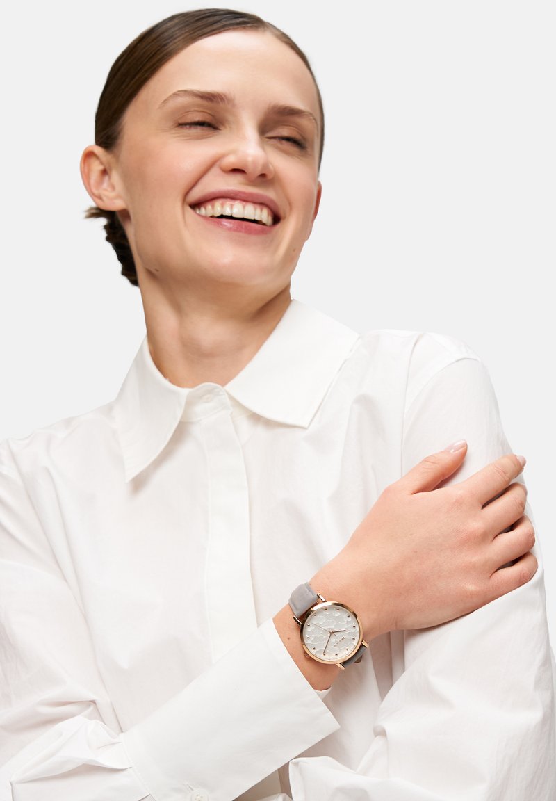 Runde Uhr mit einem strukturierten weißen Ziffernblatt und goldenen Akzenten, ausgestattet mit einem weichen grauen Armband. Am Handgelenk getragen, kombiniert mit einem eleganten weißen Hemd.