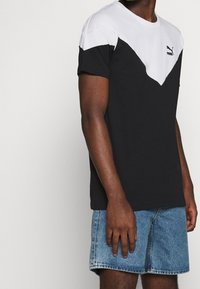 Muž oblečený v černobílém tričku značky Puma a modrých džínových šortkách, stojící před jednobarevným světlým pozadím.