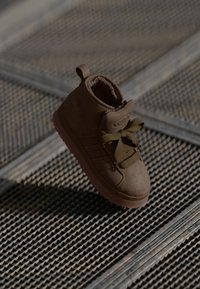 Brune semskede høy-top sneakers med en teksturert gummisåle, snørebånd, en polstret krage og en logo på tungen, vist mot en teksturert overflate.