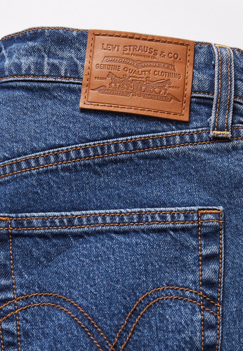Jean en denim avec une patch en cuir beige affichant "Levi Strauss & Co." Design riveté, couture contrastante orange et détails de poche arrière.