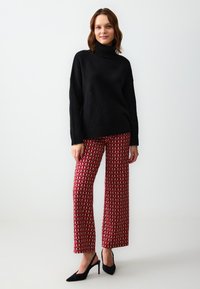 Maglione nero a collo alto, in maglia spessa. Pantaloni rossi a fantasia con forme crema. Tacchi stiletto neri. Sfondo bianco. Presentazione minimalista.
