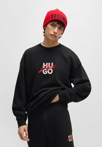 Felpa nera con logo bianco e rosso, dotata di collo a giro e vestibilità ampia. Abbinata a pantaloni neri e a un berretto rosso a costine con testo.
