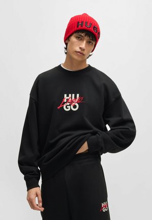 Sudadera negra con un logo blanco y rojo, con cuello redondo y corte holgado. Combinada con pantalones negros y un gorro de canalé rojo con texto.