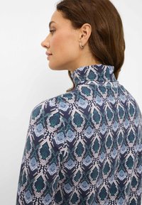 Top a maniche lunghe con motivo e colletto alto. Il tessuto presenta un design geometrico multicolore in tonalità di blu e verde su uno sfondo scuro.