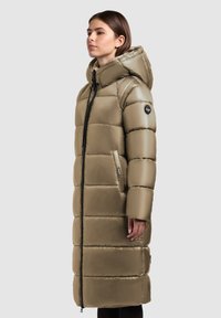 Palton lung, de culoare verde măsliniu, cu puf, cu glugă, închidere cu fermoar și două buzunare laterale. Prezintă secțiuni matlasate și un insignă de marcă pe mână.