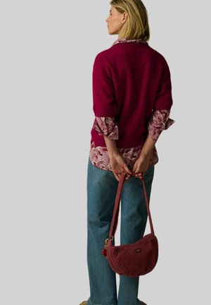 Woman facing sideways wearing a burgundy sweater over a patterned shirt, blue jeans, holding a textured burgundy handbag behind her back.