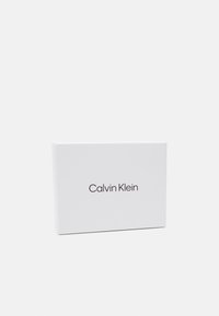 Baltas stačiakampis dėžutė su lygiu paviršiumi; "Calvin Klein" tamsiu, moderniu šriftu, centruotu ant priekinio krašto. Minimalistinis dizainas.