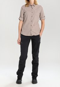 Camicia button-up beige chiaro con maniche arrotolate, abbinata a pantaloni cargo neri con tasche con zip e materiale resistente. Le scarpe nere completano l'outfit.
