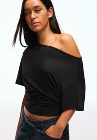 Fekete vállra húzható, puha anyagból készült felső, rövid ujjal és testhez simuló fazonnal. Sötét, lazább fazonú nadrággal párosítva.