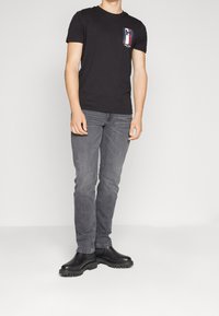 Camiseta negra de manga corta con un logo de color en el lado izquierdo del pecho; combinada con unos vaqueros grises y botas negras de suela gruesa.