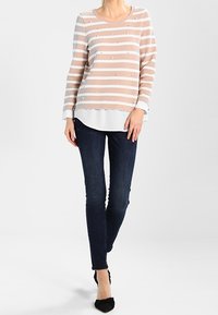 Haut léger rayé beige et blanc avec des clous. Il présente des manches longues, un col rond et un ourlet blanc en couches. Porté avec un jean foncé.