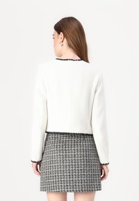 Veste blanche texturée avec des bordures noires au col et aux poignets, assortie à une jupe courte à motifs à carreaux noir et blanc.