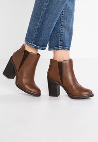 Stivali alla caviglia in pelle marrone con pannelli laterali elastici, tacchi a blocco alti, punte arrotondate e suole nere texturizzate, indossati con jeans blu risvoltati.